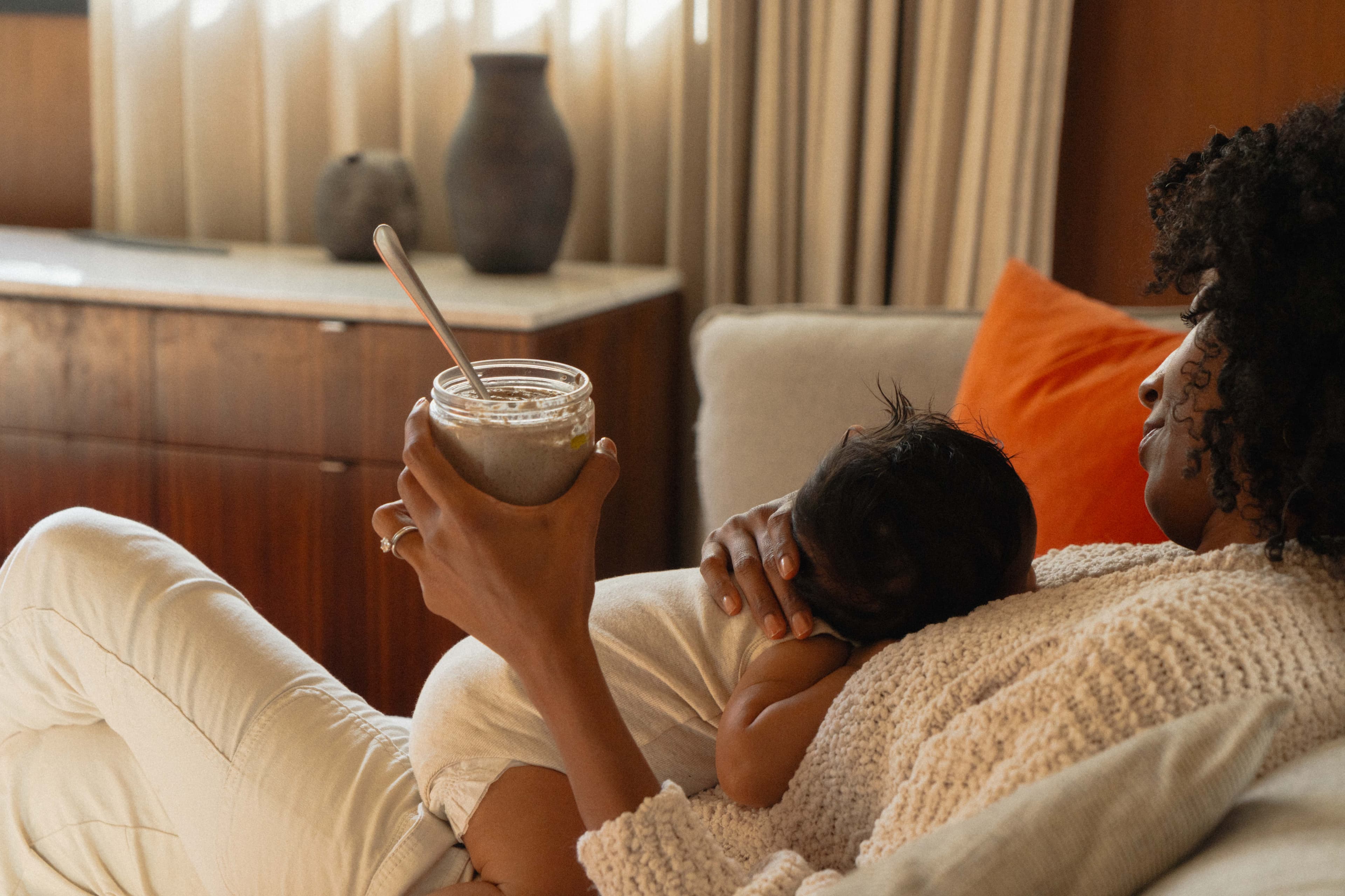 A mother resting on a sofa with her newborn, holding a Milky Oat jar